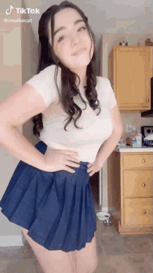 a woman in a white shirt and blue pleated skirt is standing in a kitchen .