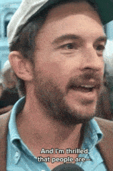a man with a beard is talking into a microphone and says and i 'm thrilled that people are ..