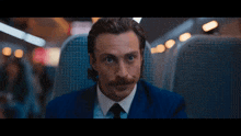 a man with a mustache wearing a suit and tie looks at the camera