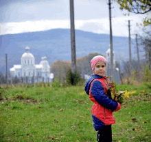 a little girl wearing a pink hat and a red jacket is holding leaves in a field