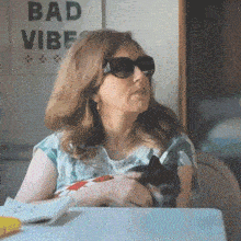 a woman wearing sunglasses sits at a table with a cat in front of a sign that says " bad vibe "