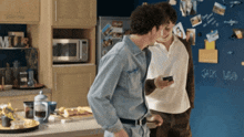 two men are standing in a kitchen and one is looking at a cell phone