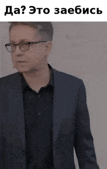 a man in a suit and glasses is standing in front of a white wall and talking into a microphone .