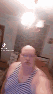a bald man wearing glasses and a striped tank top is standing in a room with a light hanging from the ceiling .