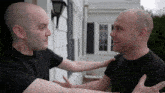 two bald men in black shirts are hugging each other in front of a house