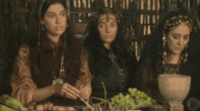 three women are sitting at a table with a bowl of grapes and vegetables
