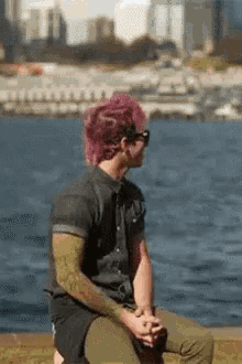 a man with pink hair is sitting in front of a body of water with a city in the background