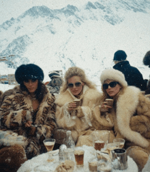 three women wearing fur coats are sitting at a table with drinks