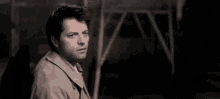 a man in a trench coat and tie is standing in a dark room and looking at the camera .