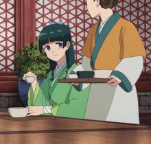 a woman in a green kimono sits at a table while a man holds a tray of cups