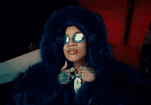 a woman wearing a blue fur coat and sunglasses has a pendant with the letter l on it