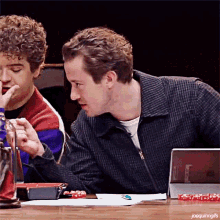 two men are sitting at a table talking to each other and one of them is holding a bottle .