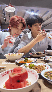 two young men are sitting at a table with bowls of food and one of the bowls has yum noodle on it