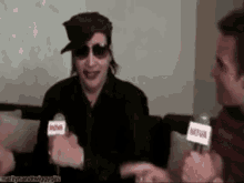 a man wearing sunglasses and a hat is talking into a microphone while sitting on a couch .