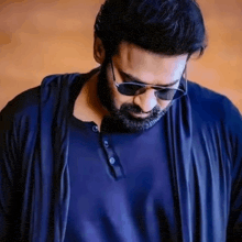 a man with a beard and sunglasses is wearing a blue shirt and a black jacket .