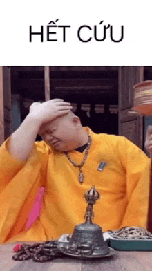 a man in a yellow robe is sitting at a table with his hand on his head and a bell in front of him .