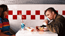 a man and a woman are sitting at a table in a restaurant eating hamburgers and drinking milkshakes .