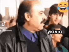 a man with a mustache is standing in front of a crowd of people and laughing .