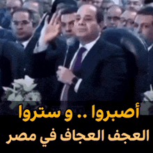 a man in a suit and tie stands in front of a crowd with arabic writing on the bottom