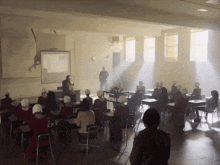 a group of people sitting in a classroom with a man standing in the background