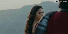 a woman in a red dress looks at a man in a black jacket in front of mountains