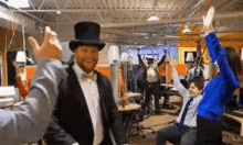 a man in a tuxedo and top hat is dancing in an office with his arms in the air .