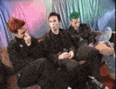 three men are sitting on a couch in a room . one of the men has green hair .