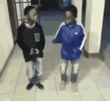 two boys are standing next to each other in a hallway and one has the letter r on his shirt .