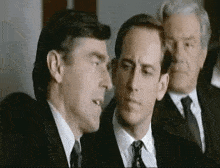 three men in suits and ties are standing next to each other in a room and talking .