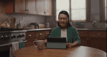 a woman sits at a kitchen table with a tablet and a cup of coffee