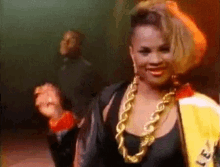 a woman wearing a gold chain necklace and a yellow jacket is dancing in a dark room .