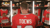 a man in a red adidas jersey with the name toews on the back stands in a locker room
