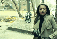a woman in a trench coat holds a gun in front of a parked motorcycle