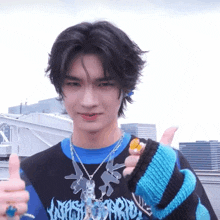 a young man wearing a sweater and a necklace giving a thumbs up gesture