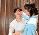 two men are standing next to each other in front of a wooden wall . one of the men is touching the face of the other .