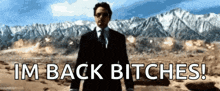 a man in a suit and tie is standing in front of mountains and says " im back bitches "