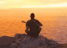 a man is sitting on a rock playing a guitar at sunset .
