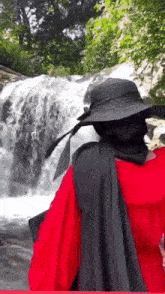 a woman wearing a black hat and a red shirt is looking at a waterfall