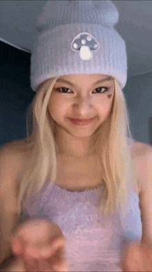 a blonde girl wearing a purple tank top and a white beanie with a mushroom on it is smiling .