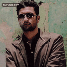 a man wearing sunglasses and a brown jacket is standing in front of a green wall .
