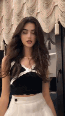 a woman in a black crop top and white skirt is standing in front of a curtain .