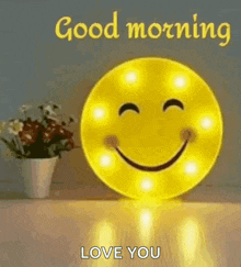 a smiley face is sitting on a table next to a vase of flowers and says `` good morning love you '' .