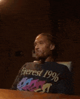 a man is sitting at a table wearing a black t-shirt that says `` everest 1996 '' .