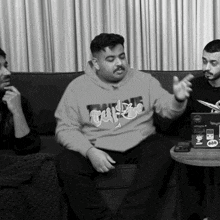 three men are sitting on a couch and one of them is wearing a hoodie that says tuf
