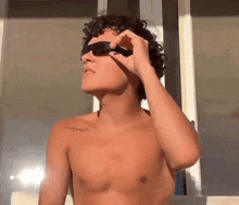 a shirtless man with curly hair is wearing sunglasses while standing in front of a window .