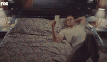 a man is laying on a bed holding a piece of paper with the fox logo behind him