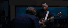 a man is playing a guitar in a dark room while another man looks on .