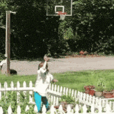 a girl stands in front of a white picket fence and a basketball hoop