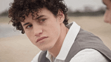 a young man with curly hair looks at the camera with a white shirt and a grey vest