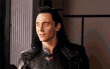 a man with long hair is wearing a black leather jacket and standing in front of a window .